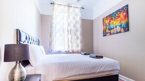 Una cama bien hecha con edredón blanco y almohadas, una lámpara de noche y un cuadro abstracto de colores en la pared.