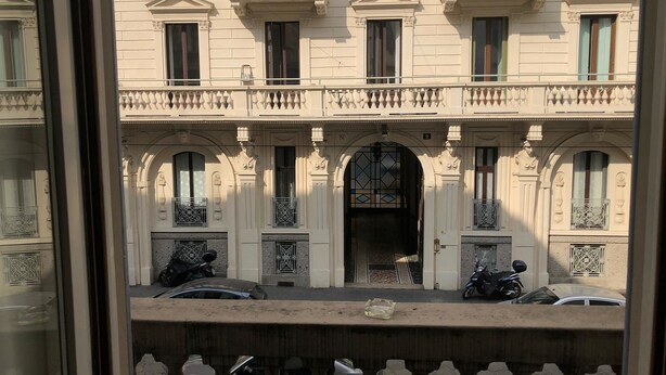 Un bâtiment à la façade symétrique, doté de nombreuses fenêtres et d’une entrée centrale surmontée d’un arc décoratif. On peut voir des voitures et une moto garées devant.