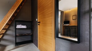 A modern interior with a wooden staircase, a black shelf with towels, and a mirror reflecting a room with a TV and a framed picture. - VILLA BRICOLA - Boutique Apartments (Karlovy Vary)