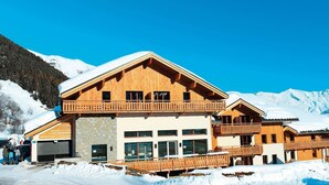 Una cabaña de madera con área de recepción y estacionamiento, rodeada de nieve y montañas.