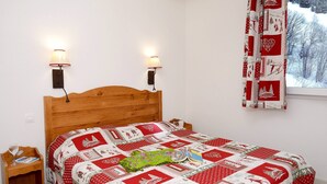 Un dormitorio con cabecera de madera, dos lámparas fijadas a la pared, colcha acolchada, manta verde y una ventana con vista a un paisaje nevado.