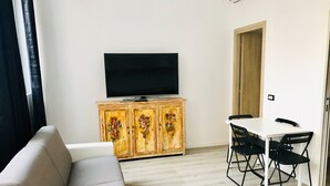 A living room with a sofa, a dining table with chairs, a TV on a wooden cabinet, and a door. - Humanitas (Milan)