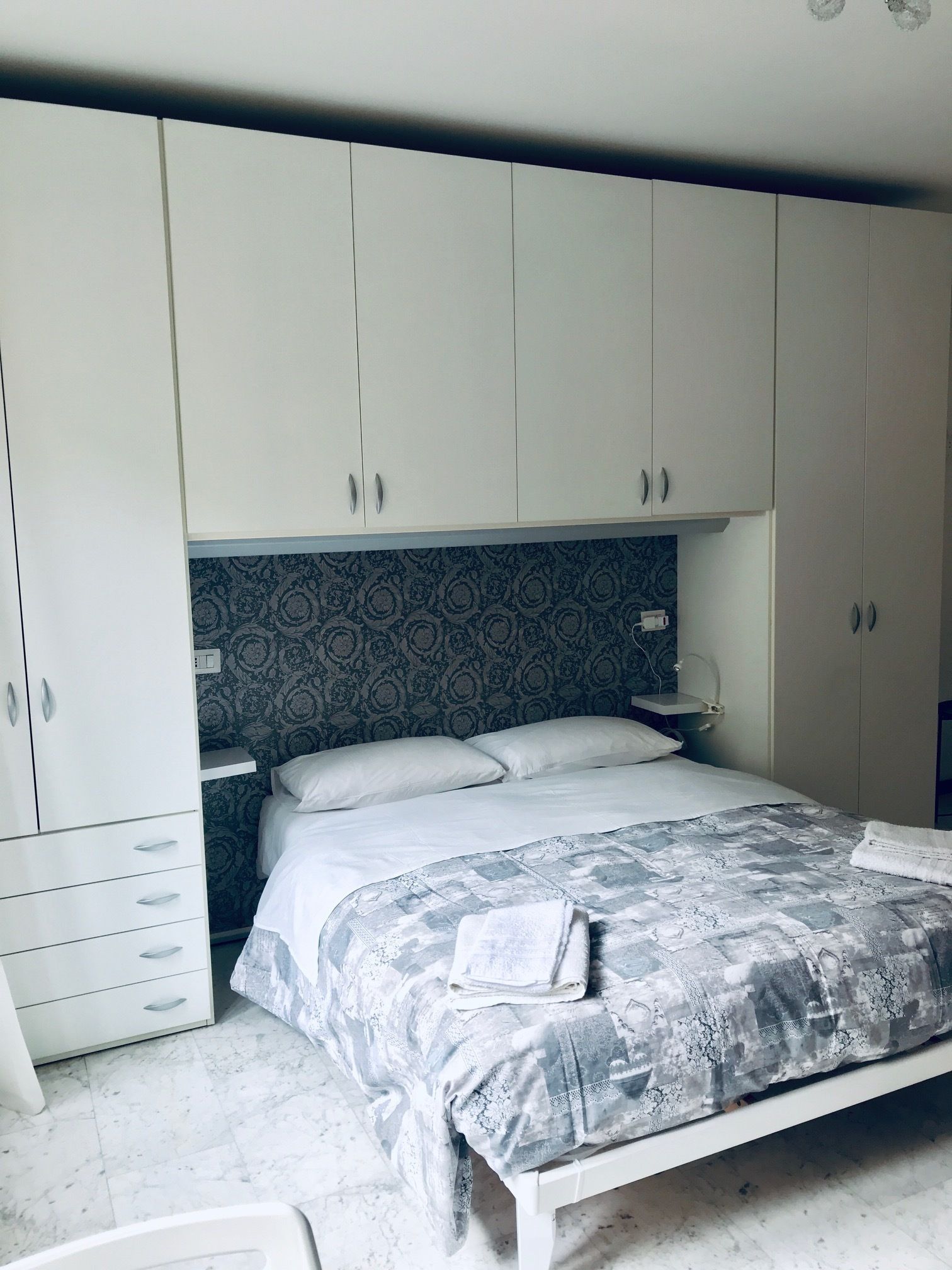 A bedroom with a bed, white wardrobe, patterned wallpaper, and a nightstand.