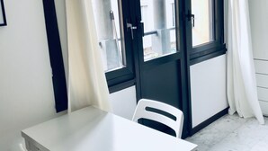 Een kamer met een witte tafel en stoel, een raam met donker frame en een witte gordijn.