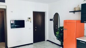 Een kamer met een flatscreen televisie aan de muur, een deur met een ronde spiegel, en een oranje kast met een plant.