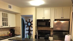 Una cucina con mobili bianchi, un piano di lavoro nero, un frigorifero in acciaio inossidabile, un forno a microonde e un fornello. Sono presenti tre sgabelli neri e una sedia di legno.