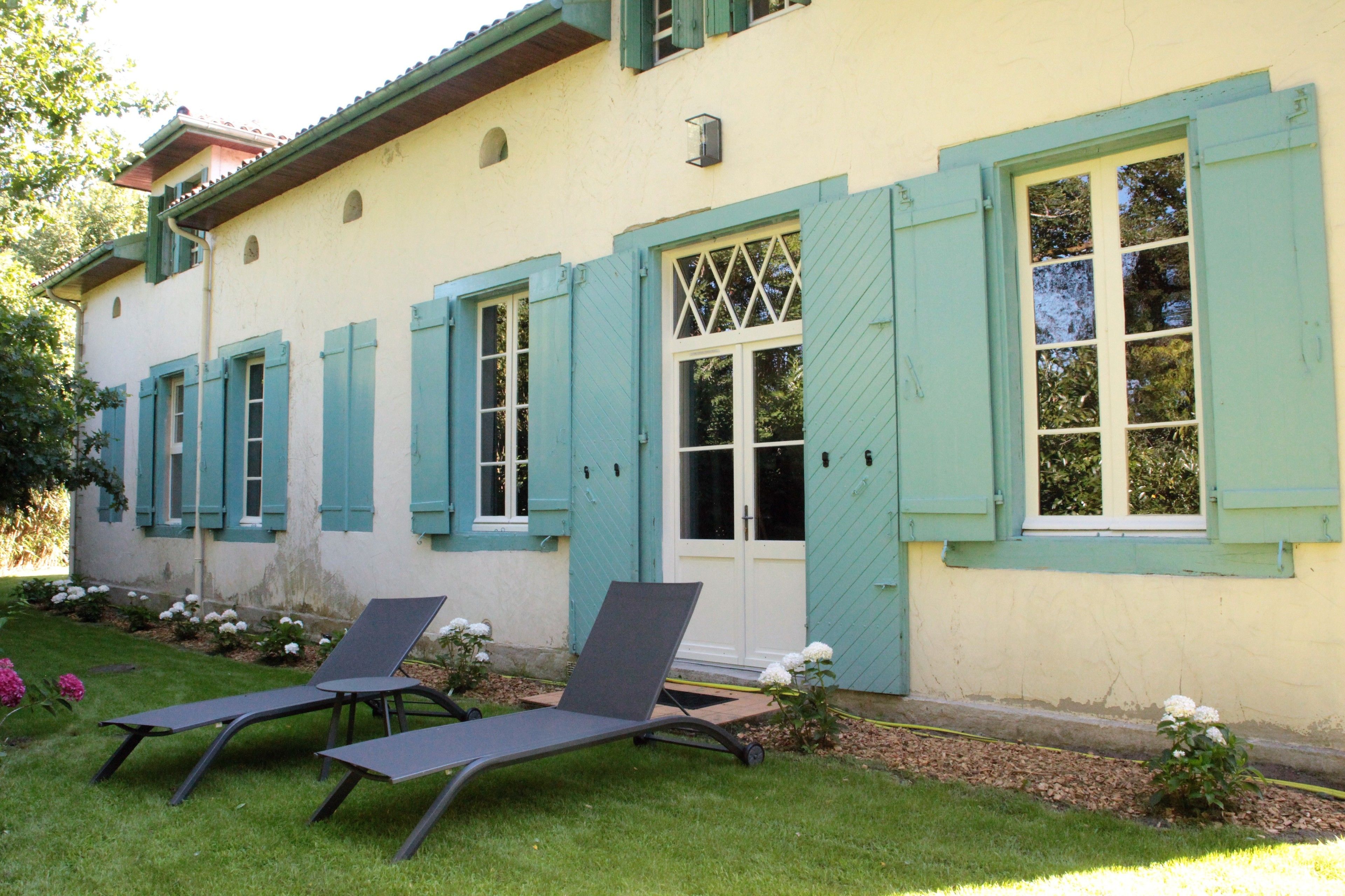 Un edificio de fachada clara y contraventanas color turquesa, con dos sillones de descanso al frente.