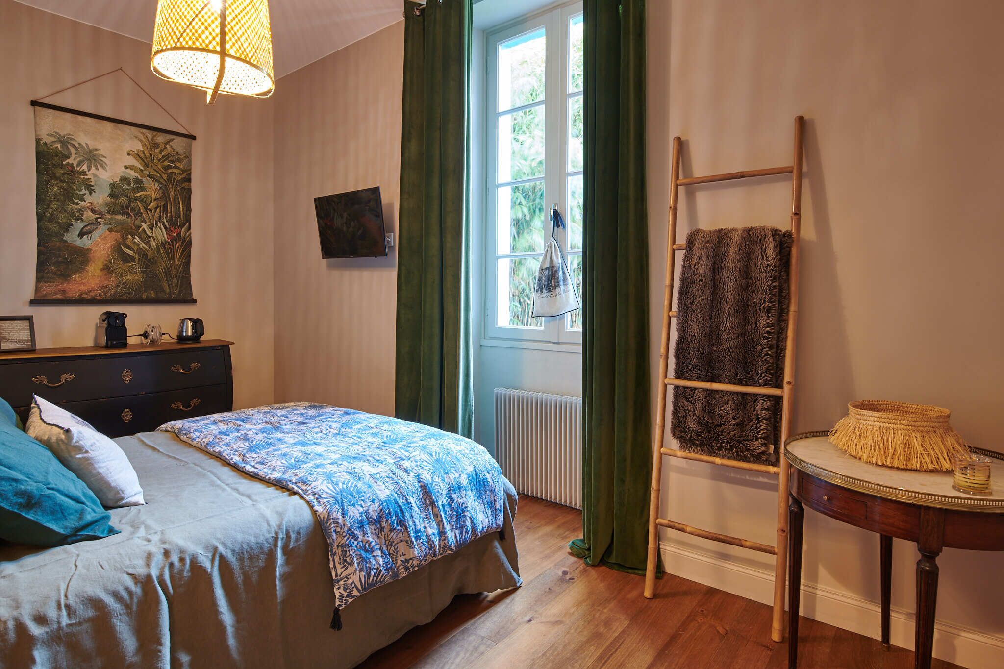 Un dormitorio con cama, cómoda, cuadro, ventana con cortinas, radiador, escalera y una mesita con una cesta.