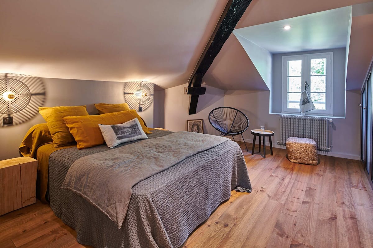 A bedroom with a large bed, a small table, a chair, a radiator, and a window with a view.
