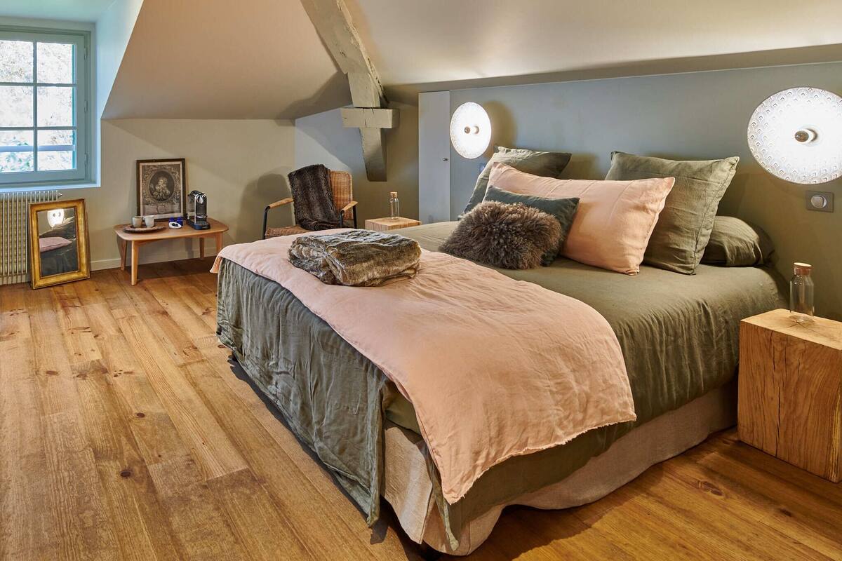 A cozy bedroom with a large bed, a wooden floor, a small table with a chair, and a window with a view.