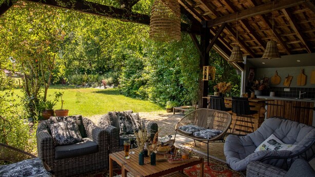 A cozy outdoor seating area with wicker furniture and a wooden table, surrounded by lush greenery and a thatched roof structure. - Bel Air Maison D'hôtes des Landes (Castets)