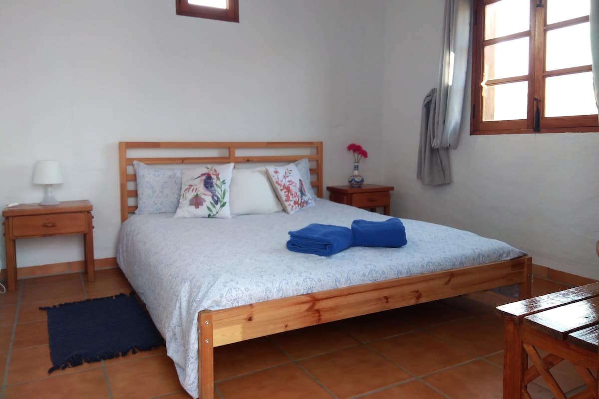 A bedroom with a wooden bed, a nightstand with a lamp, a small table, and a window with curtains.