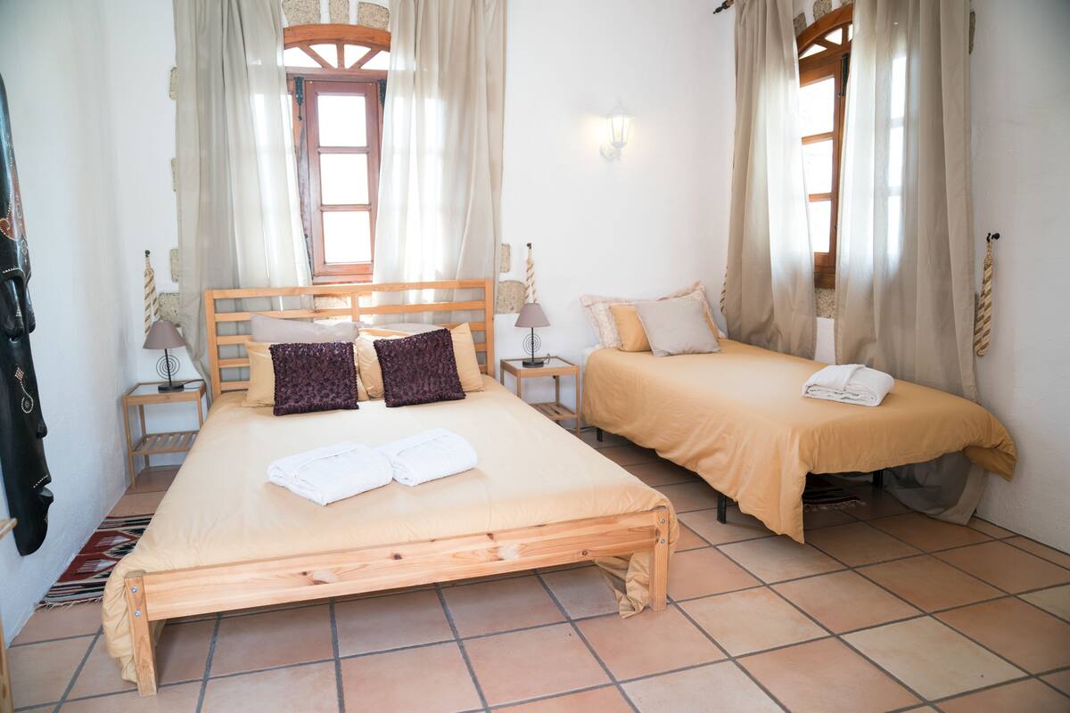 A double bed with a wooden frame, two bedside tables with lamps, a window with curtains, and a small bedside table with a lamp.