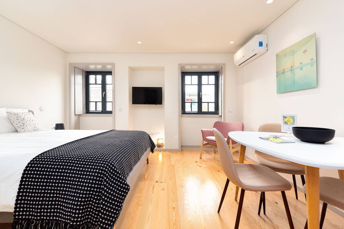 A modern hotel room with a bed, a dining table with chairs, a TV, and a painting on the wall.