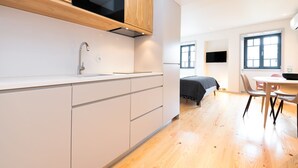 A modern kitchen with wooden cabinets, a built-in oven, and a sink. There is a dining area with a table and chairs, and a bed in the background. - Bom JardimApartments (Porto)