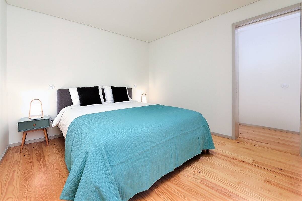 A bedroom with a bed, a nightstand, a bedside lamp, and a wooden floor.