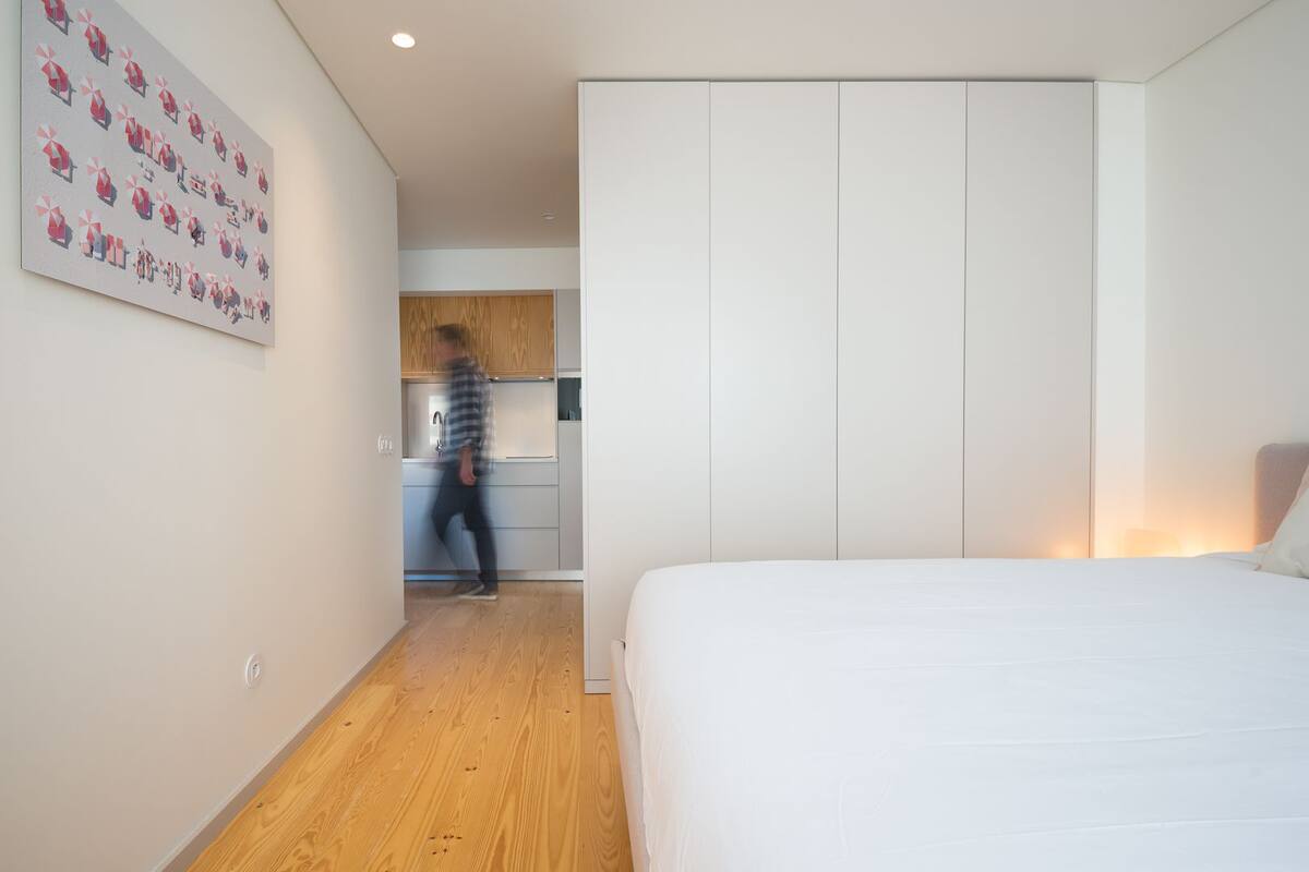 A bedroom with a large bed, a wardrobe, and a kitchen in the background.