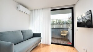 A modern living room with a grey sofa, a wooden floor, and a sliding glass door leading to a balcony with a chair. - Bom JardimApartments (Porto)