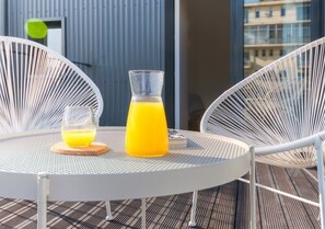 A round metal table with two white chairs, a glass of orange juice, and a bottle of orange juice on a balcony with a blue railing and a building in the background. - Bom JardimApartments (Porto)
