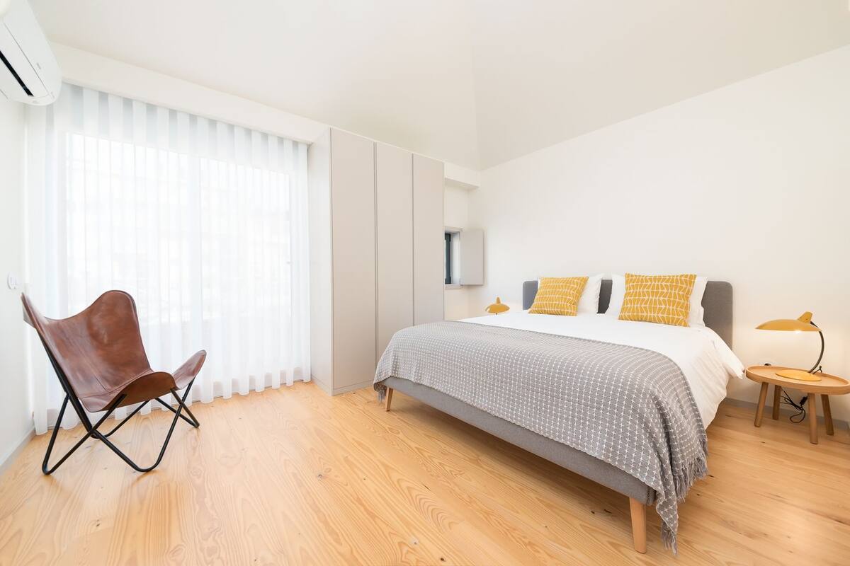 A modern bedroom with a bed, a chair, a nightstand, and a window with sheer curtains.