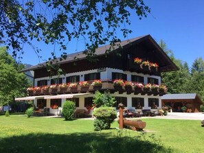 A traditional chalet with a balcony adorned with flowers, a well-maintained lawn, and a log structure in the foreground.