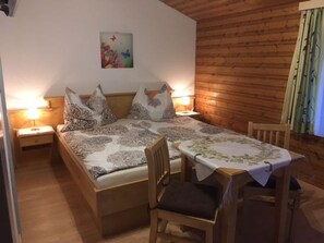 A room with a bed, two bedside tables, a chair, a table with a tablecloth, and a wooden wall.