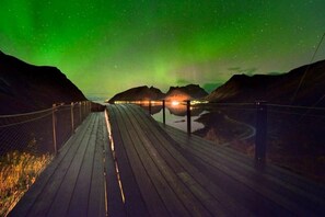 A wooden boardwalk leading to a bridge over a body of water, with a view of mountains and the Northern Lights in the background.