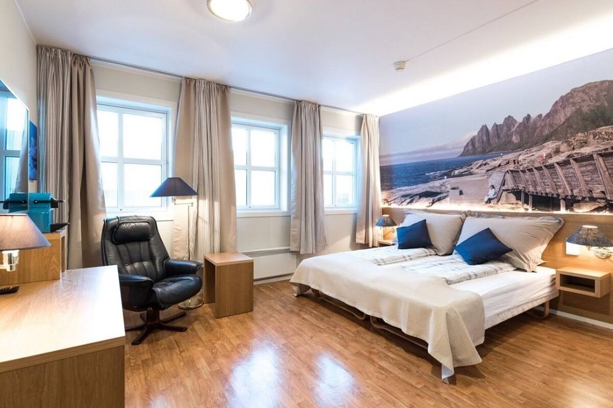 Habitación de hotel con cama, escritorio, silla, televisor y un gran ventanal con cortinas.