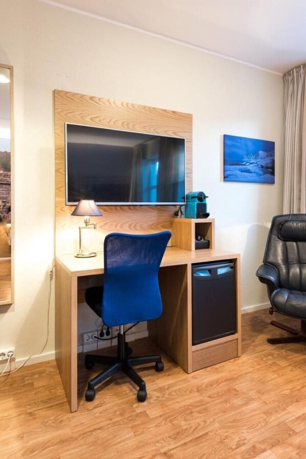 A compact room with a wooden desk, a blue office chair, a television, and a black leather chair. - Senja Hotell (Senja)