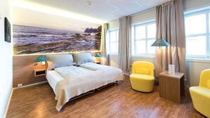 Una habitación de hotel con una cama, dos mesitas de noche, una lámpara, una silla y un ventanal con cortinas.