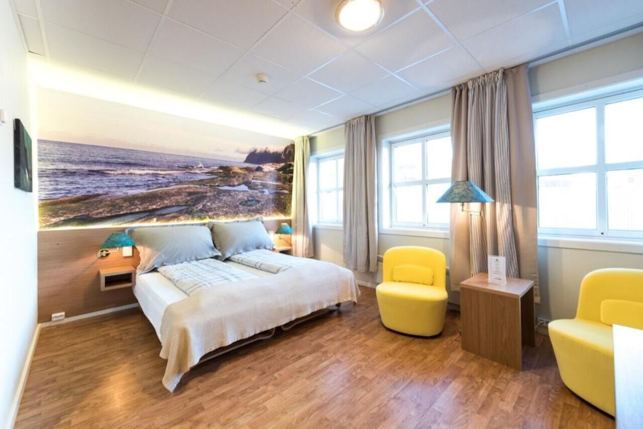 A hotel room with a bed, a yellow armchair, a small table, and a large window with curtains.