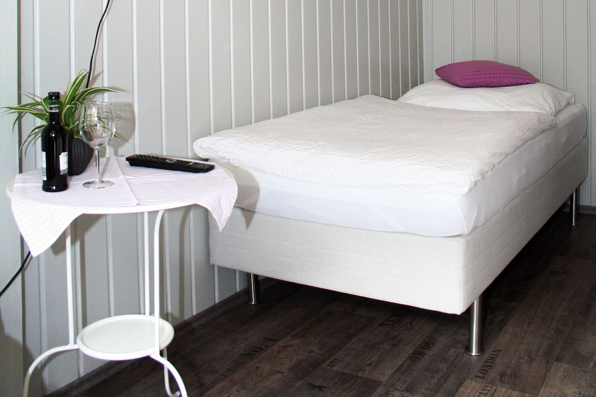 A single-bed room with a white mattress, a small bedside table with a remote control, a wine bottle, and a glass. A TV mounted on the wall displays "Hotel Klenkes".