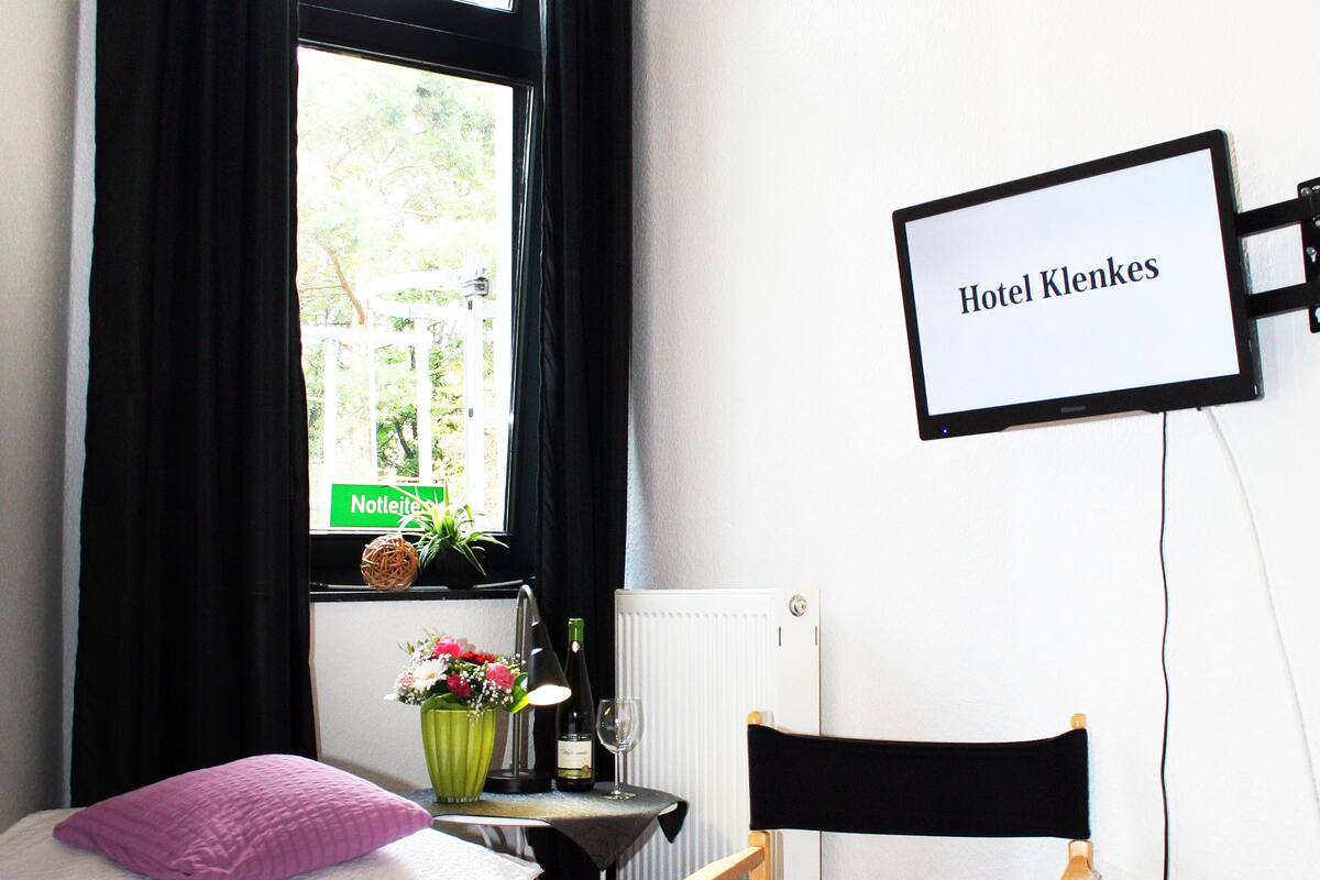 A hotel room with a bed, a desk with a computer, a chair, a window with black curtains, and a sign reading "Hotel Klernes".