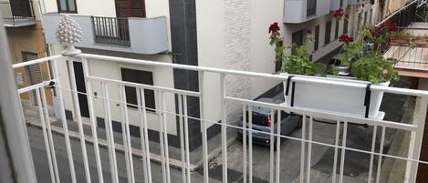 Un balcon avec une balustrade blanche, une voiture grise garée en contrebas et un pot de fleurs avec des fleurs rouges.