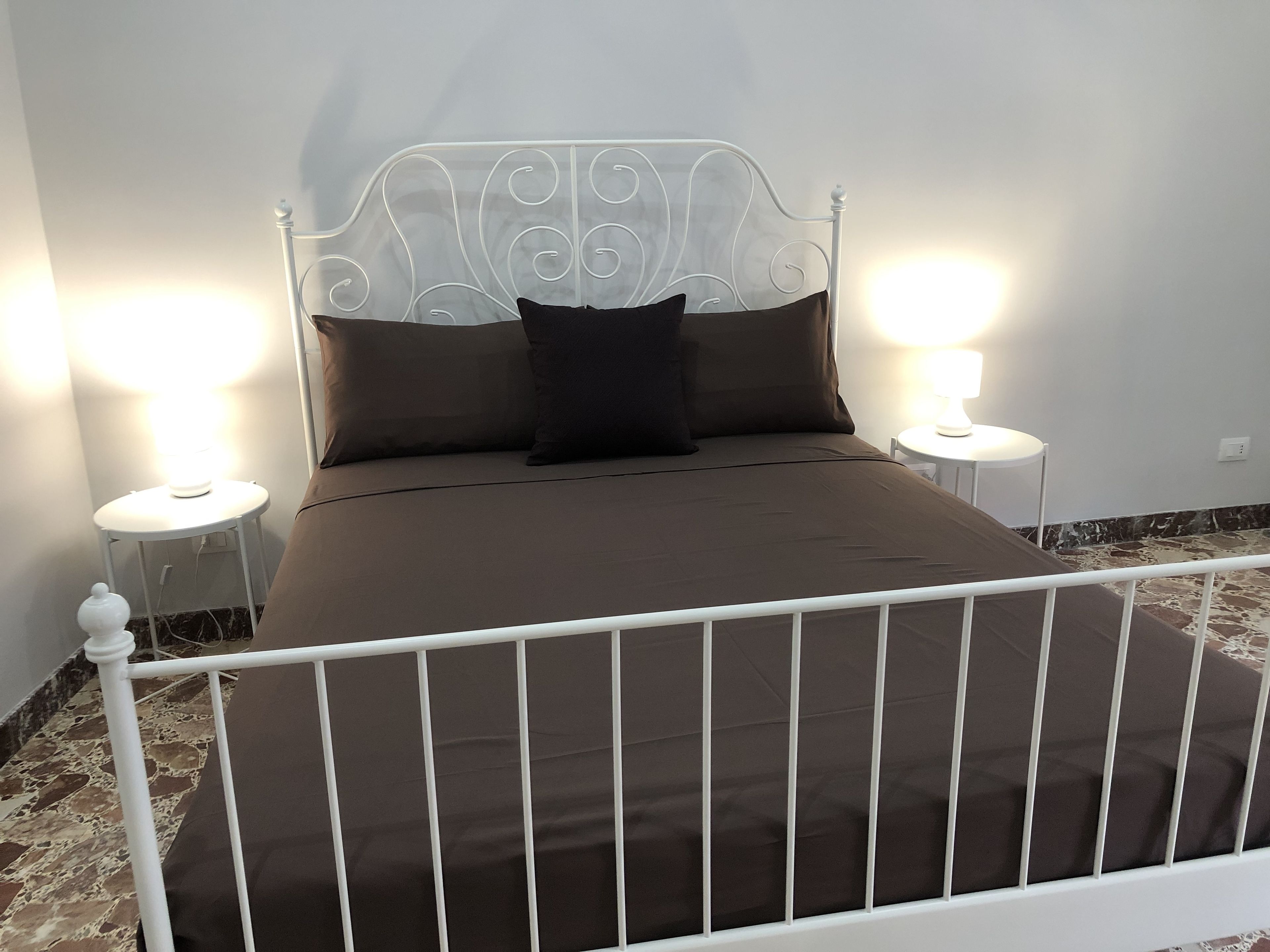 Een bed met een wit metalen frame en een bruine matras, met aan weerszijden nachtkastjes en lampen.