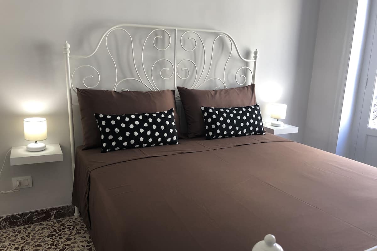 A neatly made bed with a brown bedspread and two pillows with a polka dot pattern. There are two bedside tables with lamps, and the room has a tiled floor.