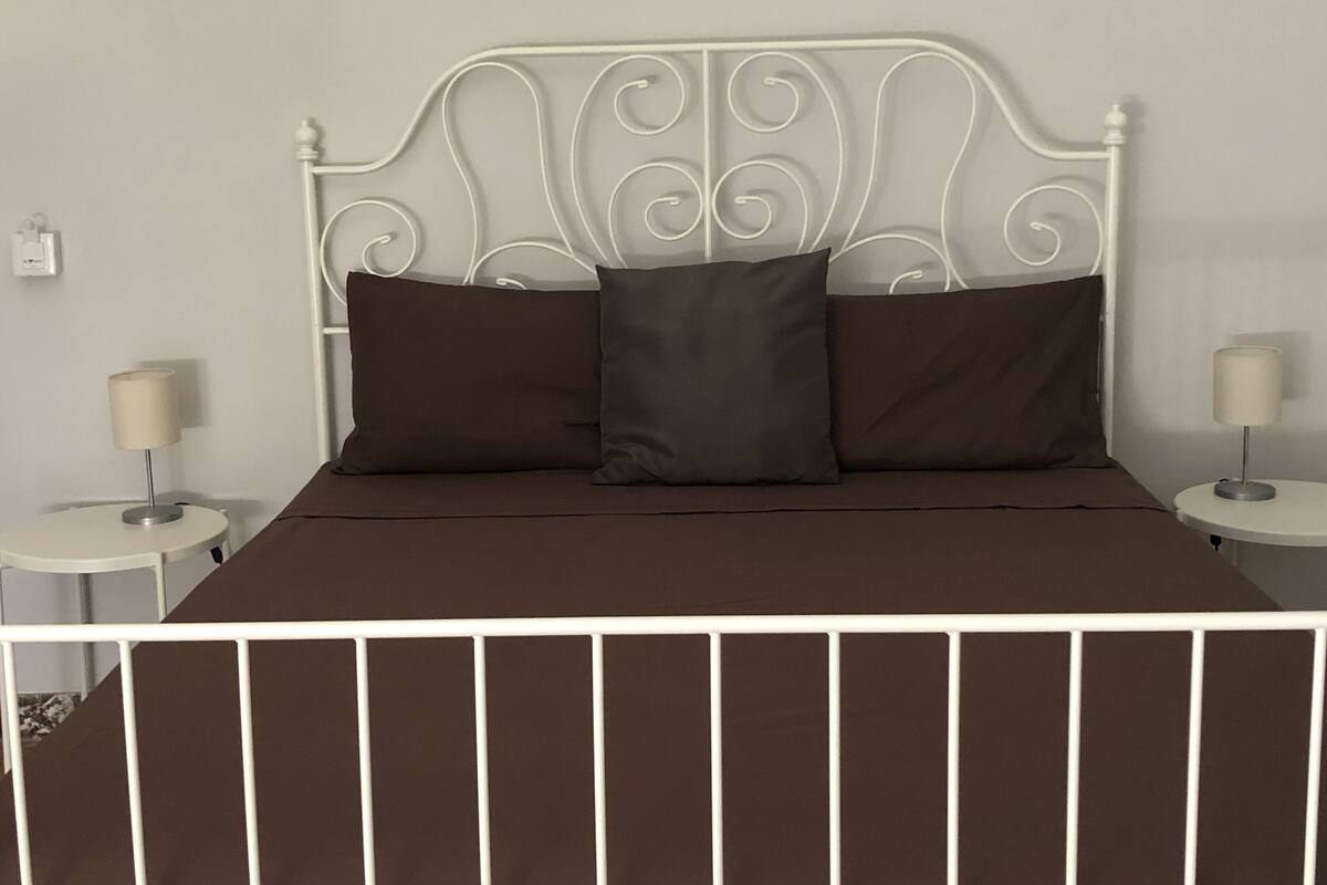 A bed with a white metal headboard, two brown pillows, and a brown bedspread. There are two bedside tables with lamps on either side of the bed.