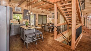 Interior de una cabaña de madera con cocina, comedor y una escalera.