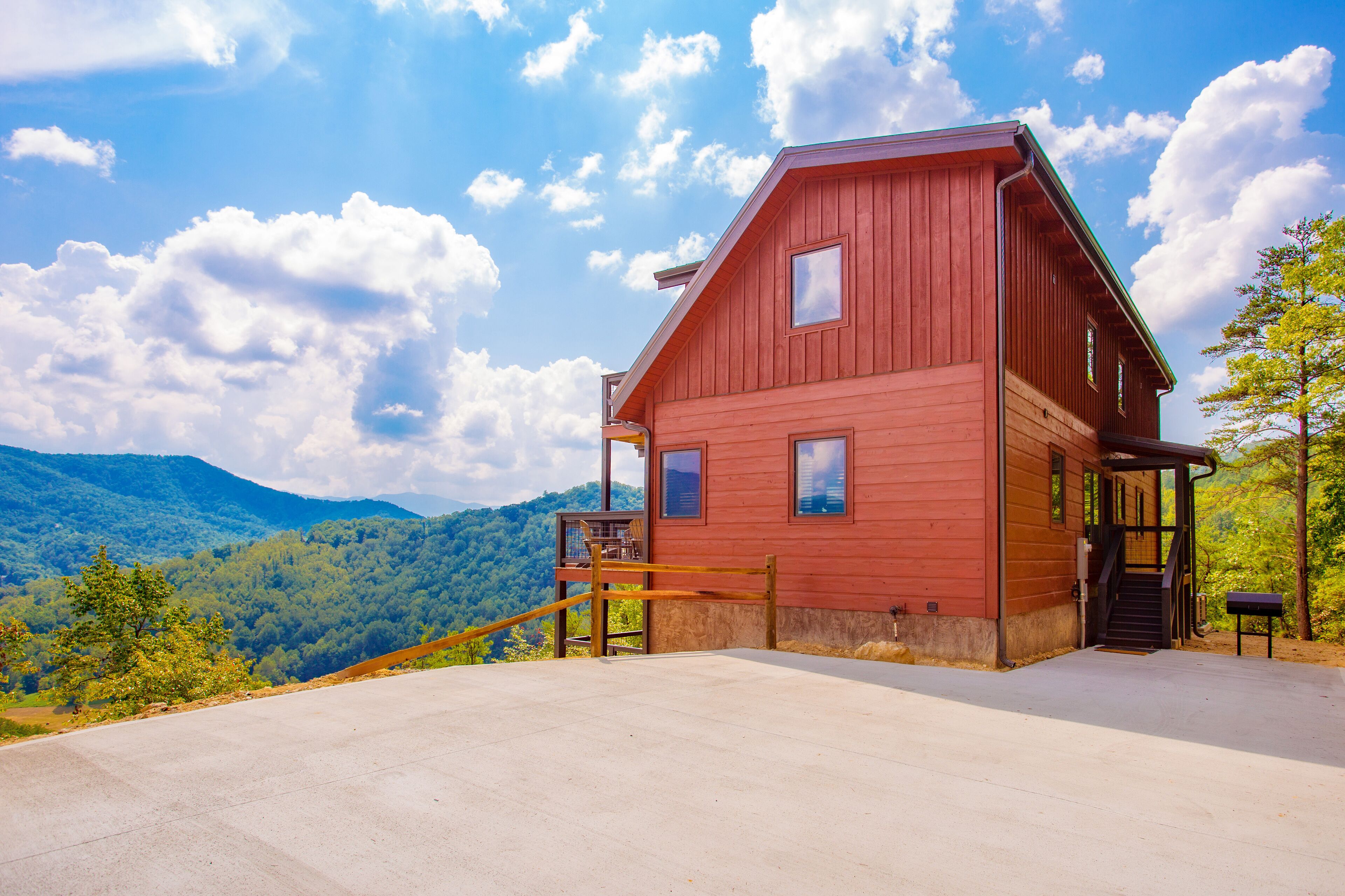 A red wooden cabin with a concrete patio, a wooden staircase, and a view of a forested mountain range.