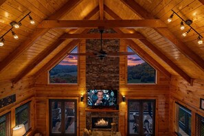 L’intérieur d’une cabane en rondins, avec une cheminée en pierre, des poutres en bois et un téléviseur.