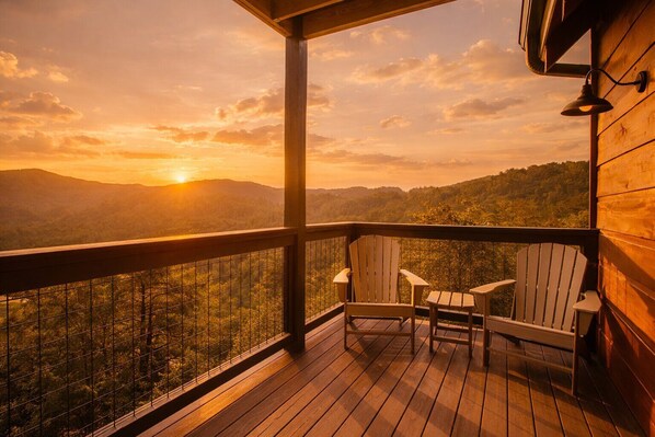 A wooden deck with Adirondack chairs, a railing, and a view of a sunset over a forested mountain range. - Amazing View Lodge- Breathtaking Views, 10 minutes to downtown Pigeon Forge! (Sevierville)