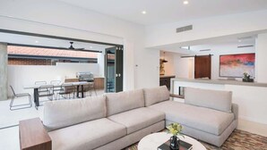 Ein modernes Wohnzimmer mit einem großen, winkelförmigen Sofa, einem runden Couchtisch und einem Essbereich. Durch eine offene Tür ist eine Küche zu sehen.