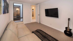 Ein Wohnzimmer mit einer Couch, einem an der Wand montierten Fernseher und einer an der Wand angelehnten Gitarre.
