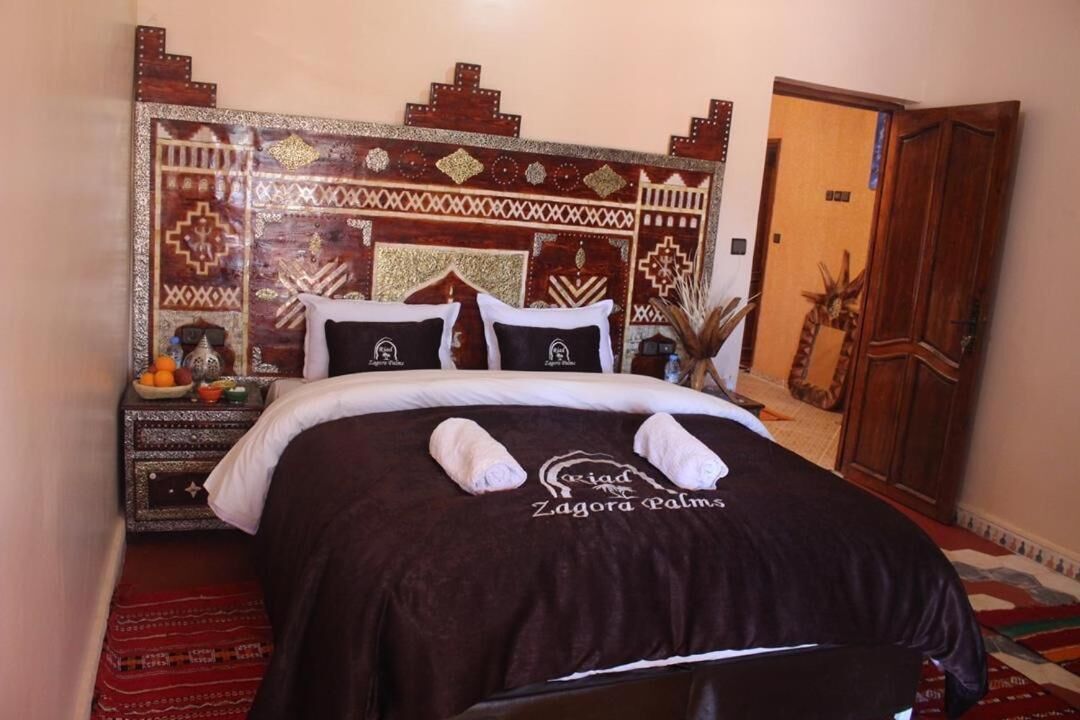 Una cama con colcha marrón oscuro, dos toallas, una mesita de noche con frutero y un cabecero decorativo.