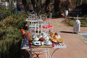 An outdoor dining table set with breakfast items, surrounded by greenery and a white metal chair.