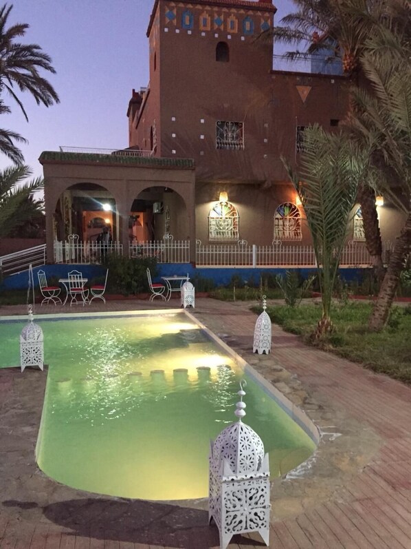Un patio con piscina, palmeras y un edificio con iluminación decorativa.