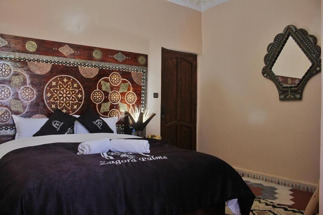Una cama bien hecha con una cubierta oscura, un cabecero decorativo y un espejo fijado a la pared.