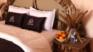 A neatly made bed with a large wooden headboard, two pillows with a logo, a small wooden nightstand with a fruit bowl and a bottle, and a decorative plant.