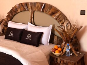 A neatly made bed with a large wooden headboard, two pillows with a logo, a small wooden nightstand with a fruit bowl and a bottle, and a decorative plant.
