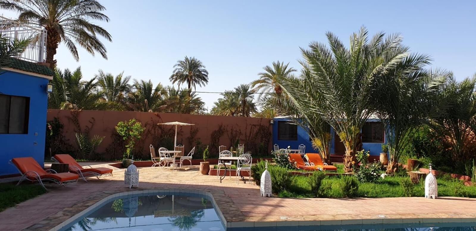 A pool area with lounge chairs, a table, and chairs, surrounded by palm trees and a blue building.